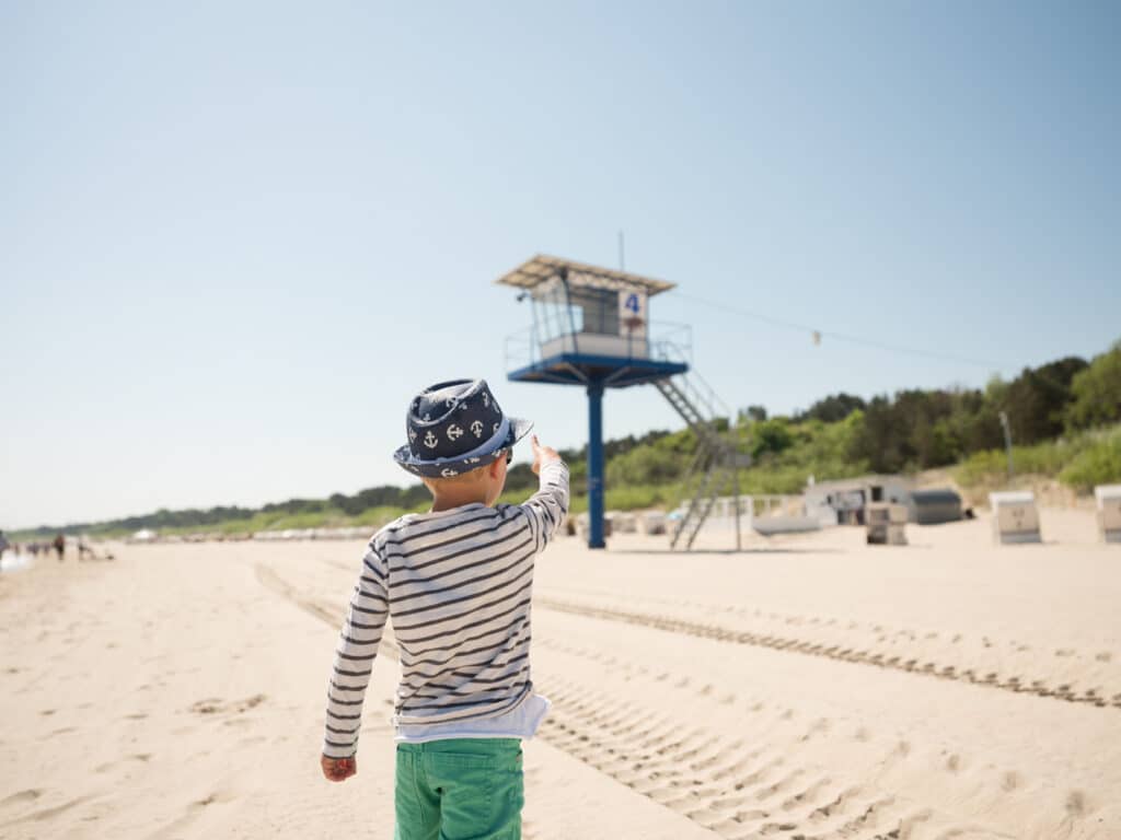 Ein kleiner Junge mit gestreiftem Pullover und blauem Sommerhut mit Ankermotiv steht auf dem feinen Sandstrand und zeigt mit dem Finger auf einen blauen Rettungsschwimmerturm.