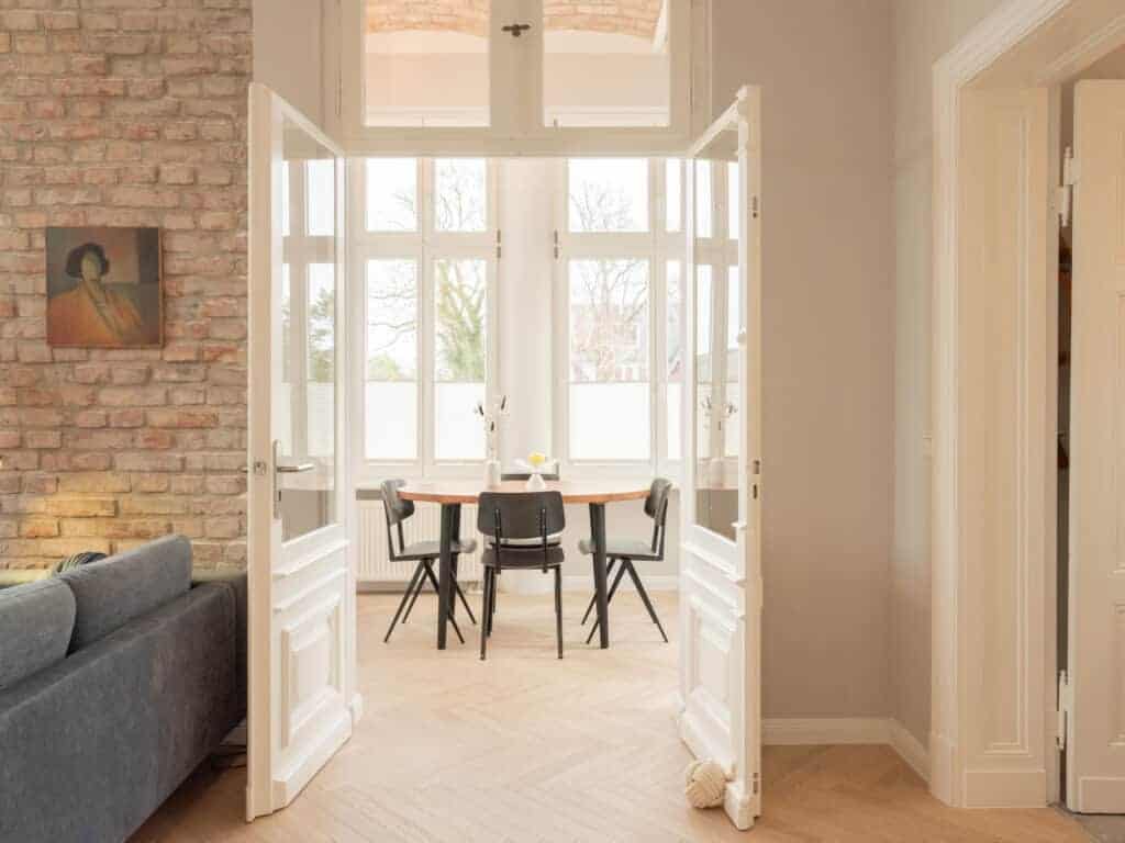 Blick durch geöffnete weiße Flügeltüren mit Oberlicht auf eine helle Loggia mit Holzesstisch, links eine Backsteinwand mit Kunstwerk.