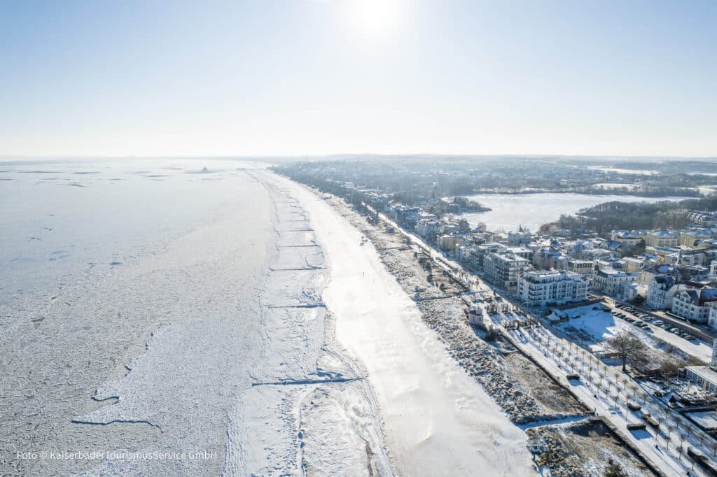 Luftaufnahme einer verschneiten Ostseeküste mit zugefrorenem Meer, weißem Sandstrand und angrenzenden historischen Bäderarchitektur-Villen unter blauem Himmel.