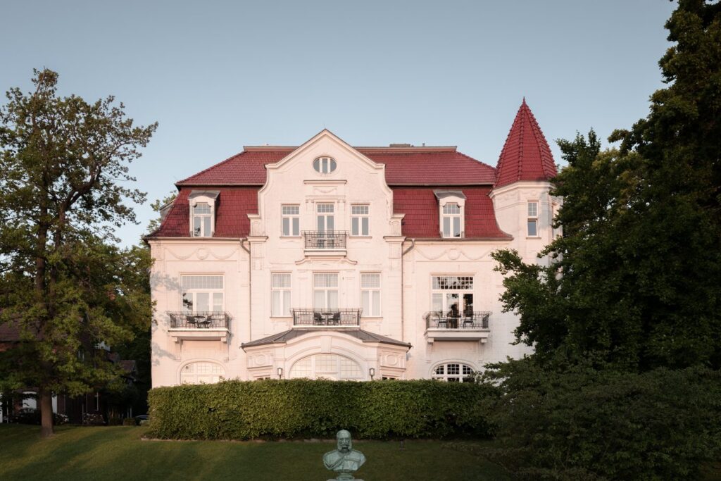 Prächtige weiße Villa der Bäderarchitektur mit rotem Ziegeldach, kleinen Erkern und Balkonen, umgeben von grünen Bäumen in der Abendsonne.