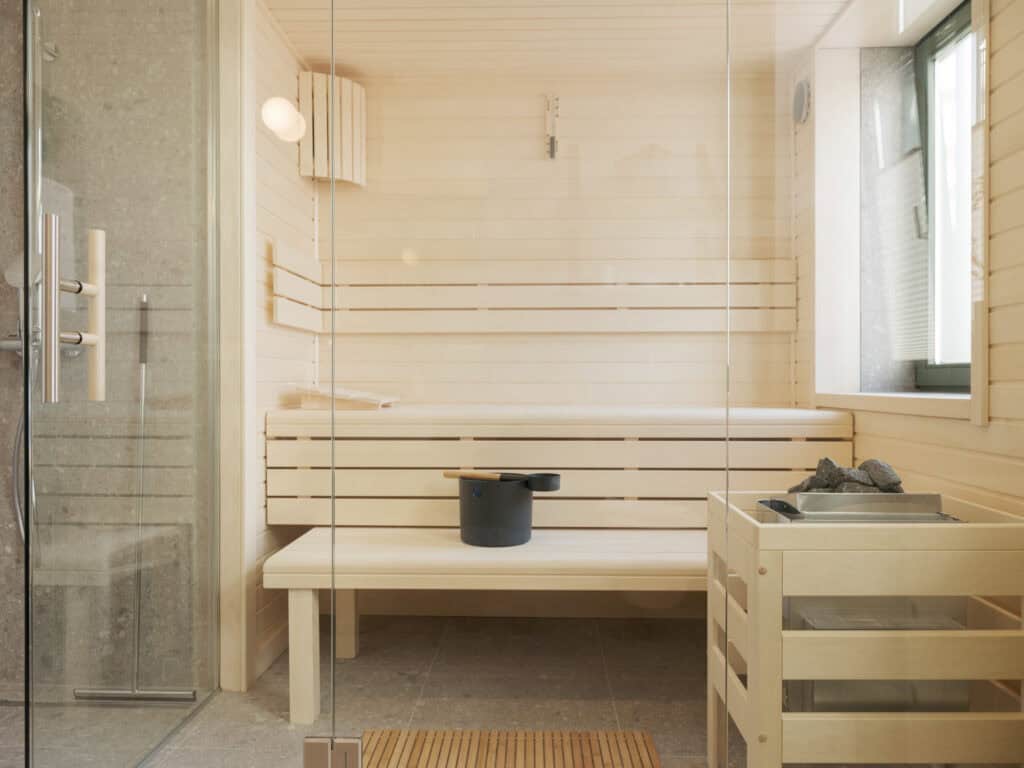 Innenansicht einer hellen Holzsauna mit zweistufiger Sitzbank, Saunaofen mit Aufgusssteinen und schwarzem Aufgusseimer.