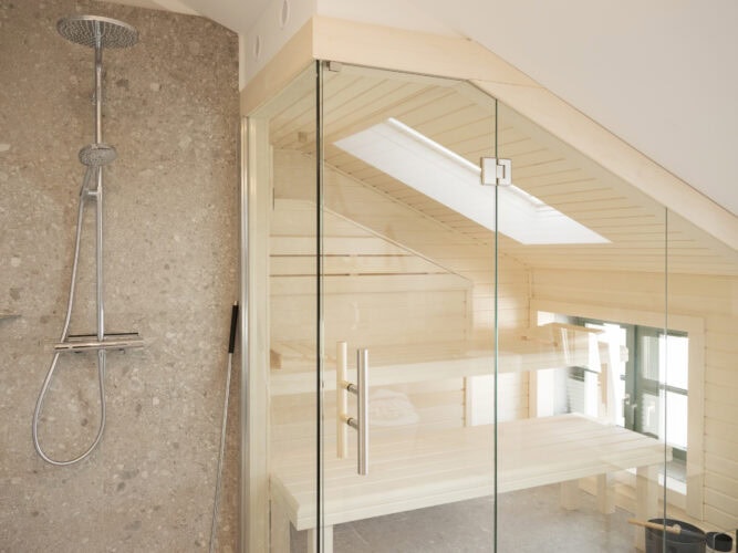 Modern gefliestes Badezimmer mit Regendusche aus Chrom und angrenzender, vollverglaster Sauna unter einem Dachflächenfenster.