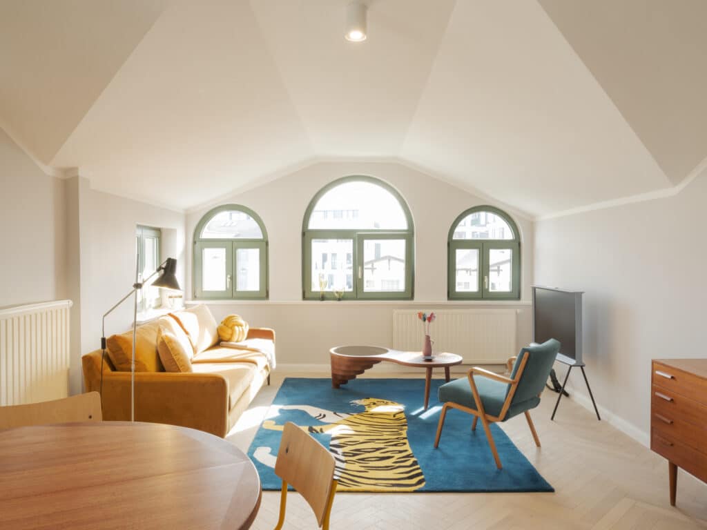 Ein helles Wohnzimmer im Dachgeschoss mit drei halbrunden Fenstern, einem gelben Samtsofa, einem markanten blauen Teppich mit Tigermotiv und einem filigranen Couchtisch aus Holz.